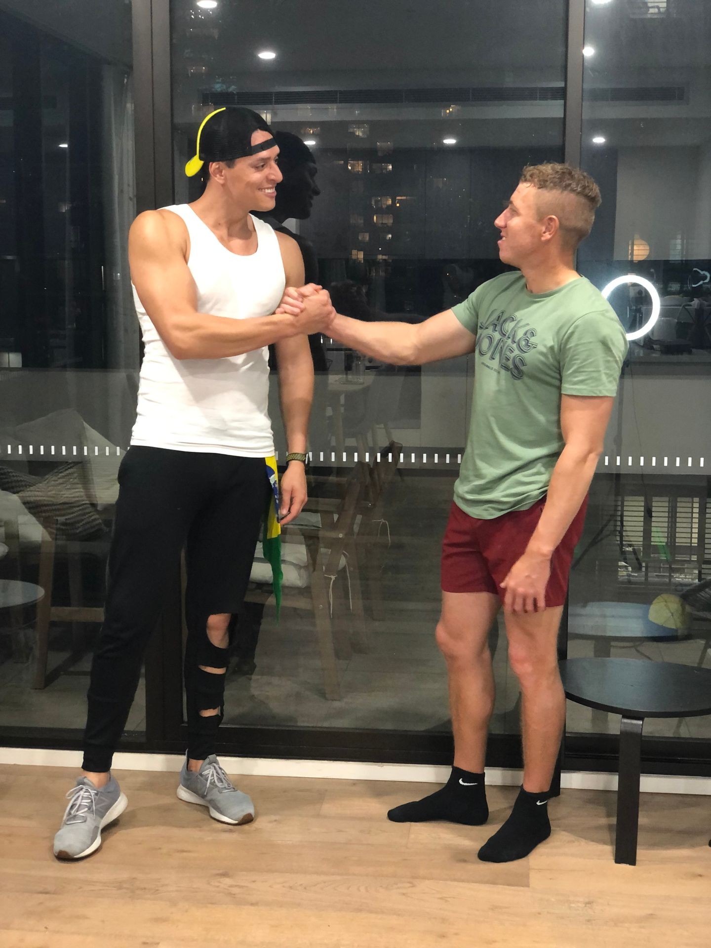 Two men wearing casual clothing shake hands and smile in front of a large window at night.