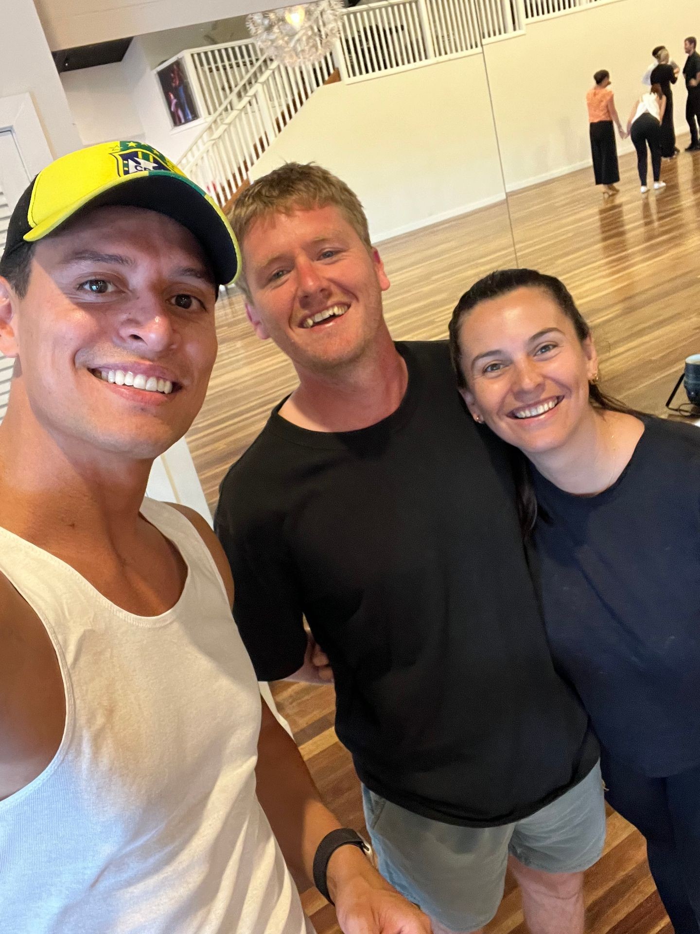 Three people standing together in a room with wooden flooring and white railings in the background.