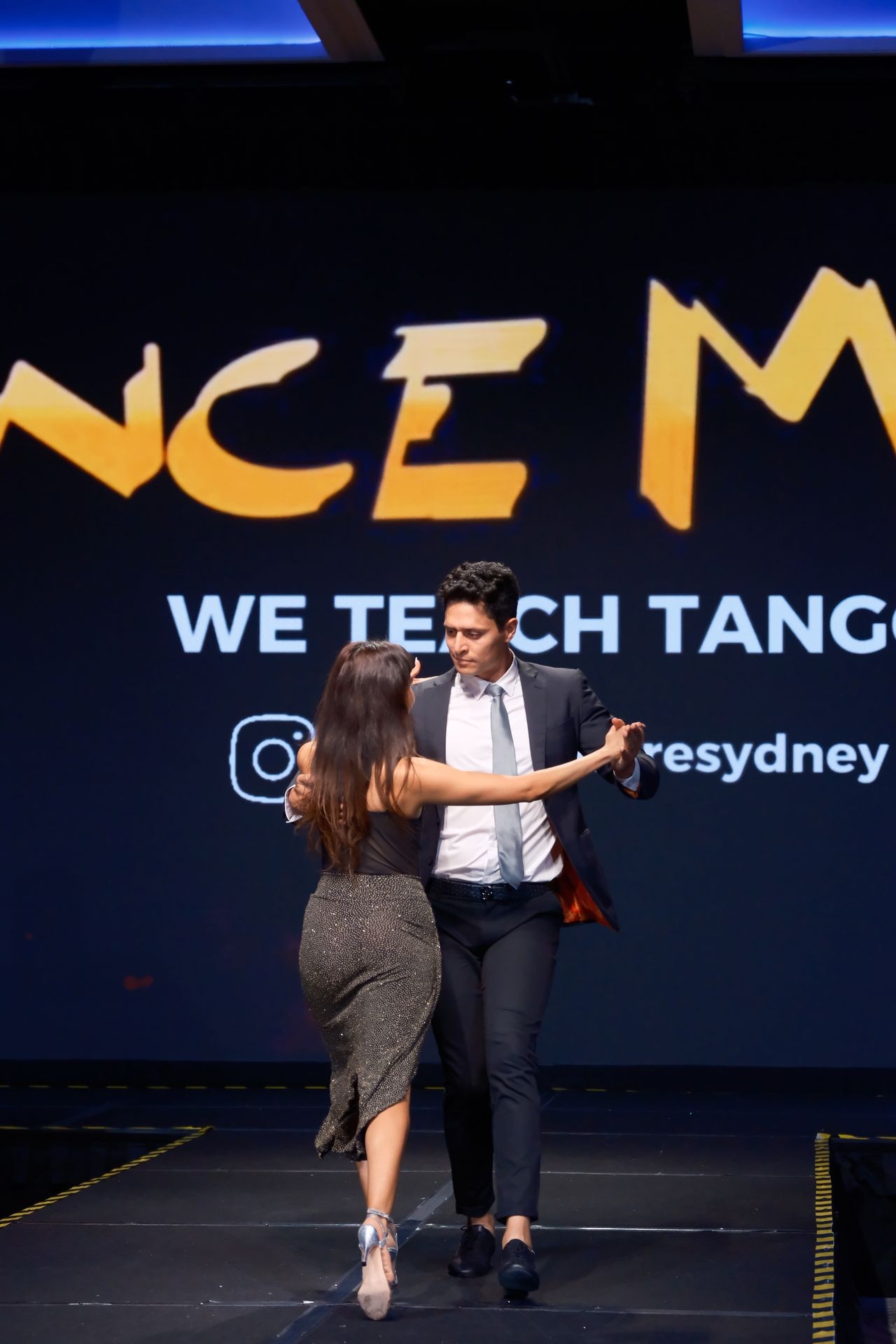 A couple performing a tango on stage with a 'We Teach Tango' sign in the background.