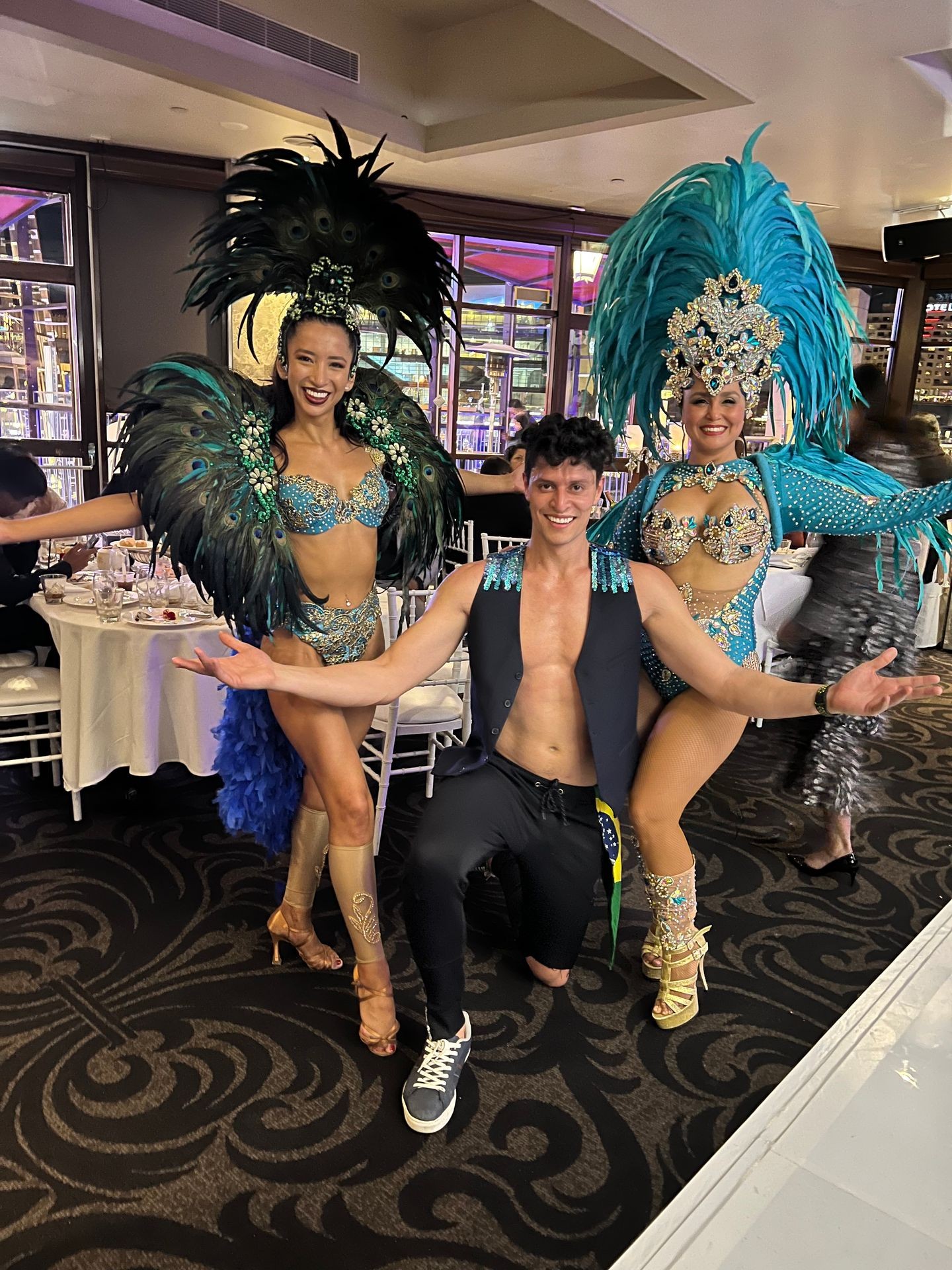 Three performers in elaborate, colorful costumes with feathered headdresses posing in an indoor event space.