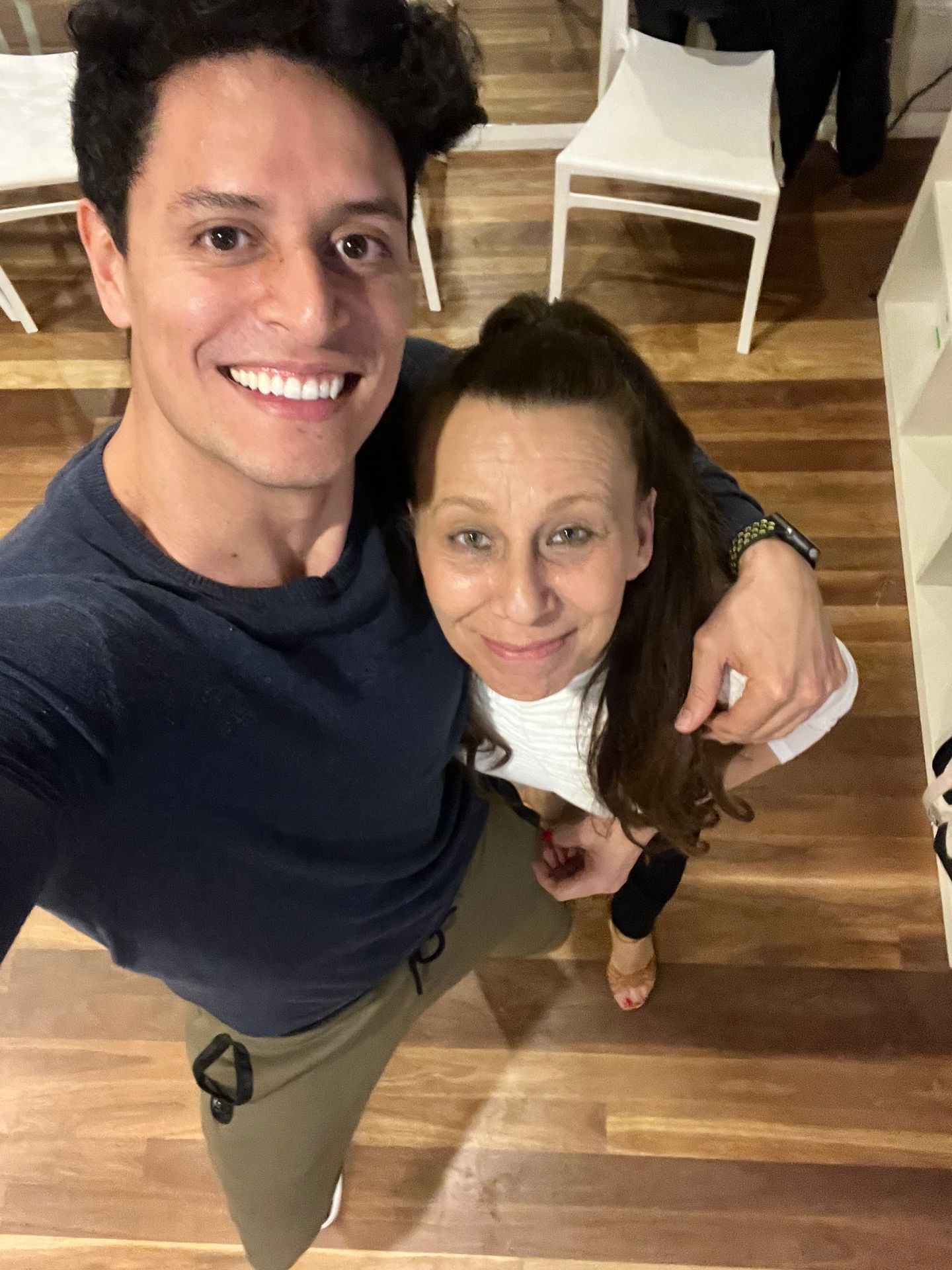 Two people taking a selfie in a room with wooden flooring and white chairs in the background.