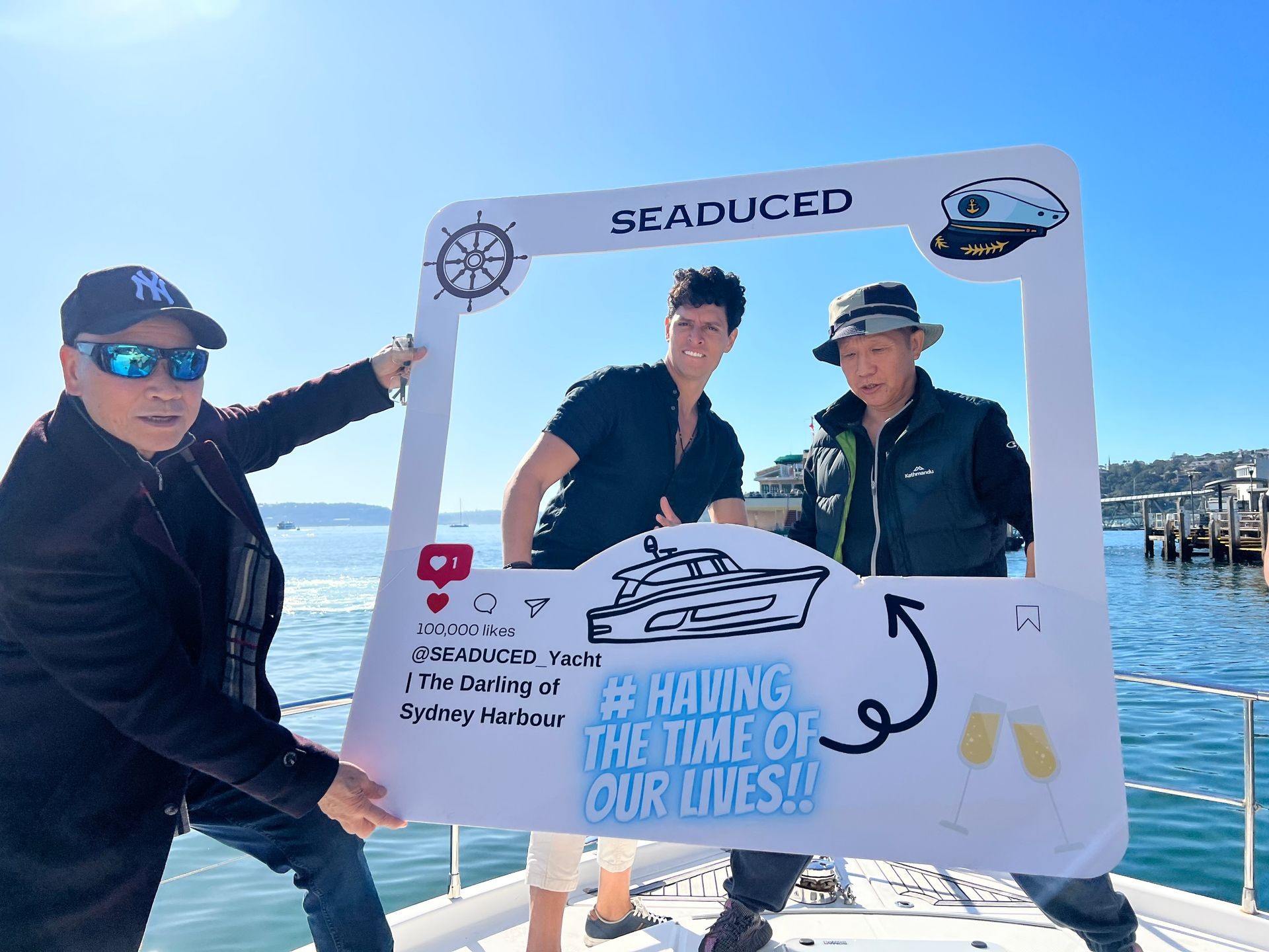 Three people holding a large photo frame prop on a boat with Sydney Harbour in the background.