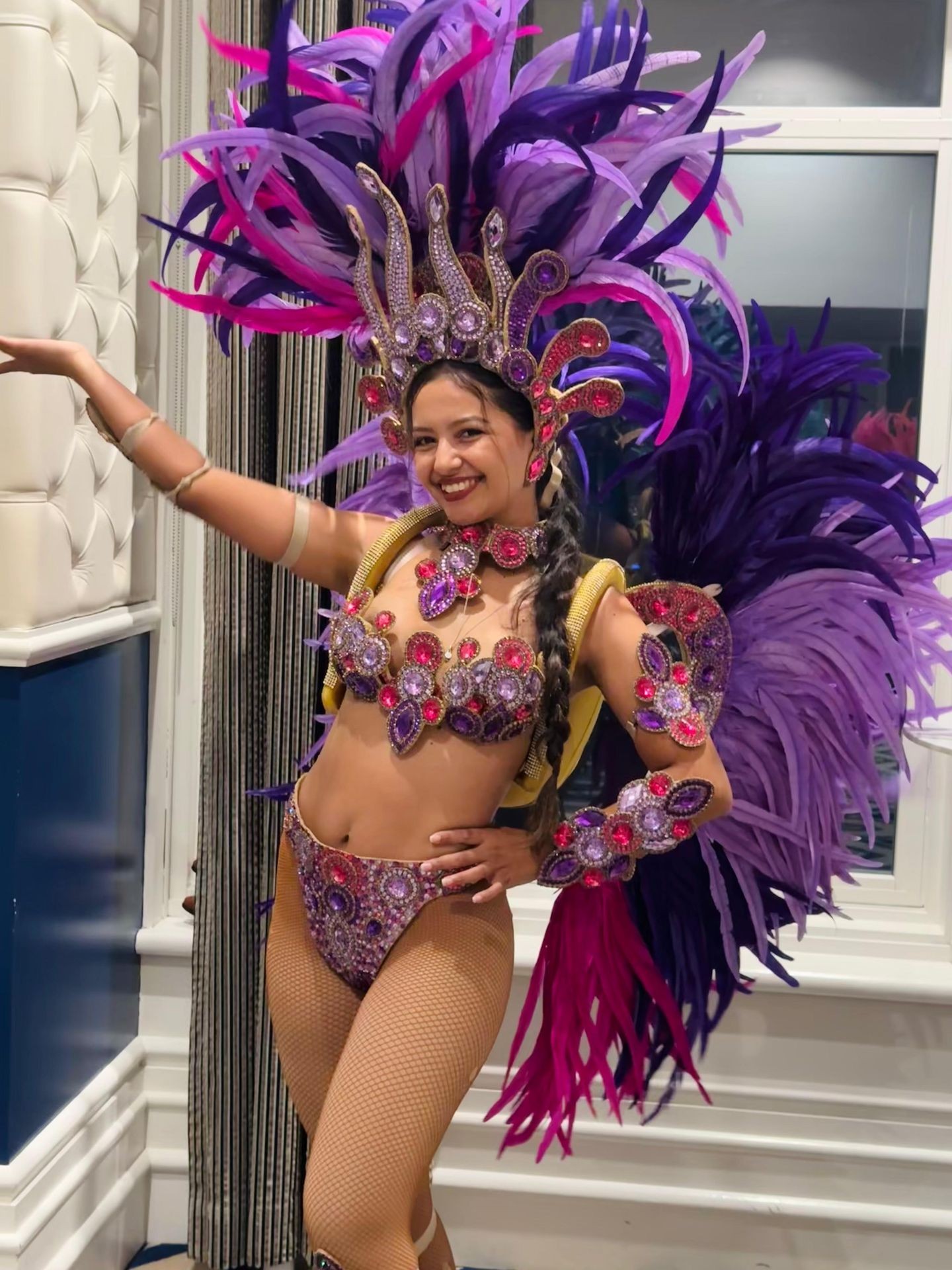 Woman in elaborate purple and pink feathered costume with jeweled accents, smiling indoors.