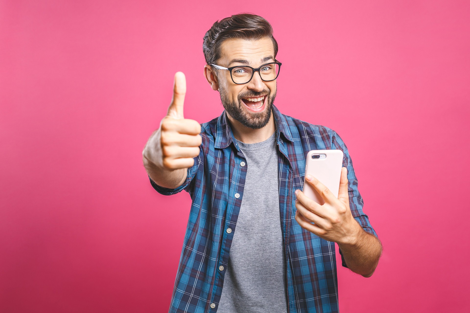 Portrait of a cheerful bearded man taking selfie and showing thumbs up gesture over pink background. Isolated.